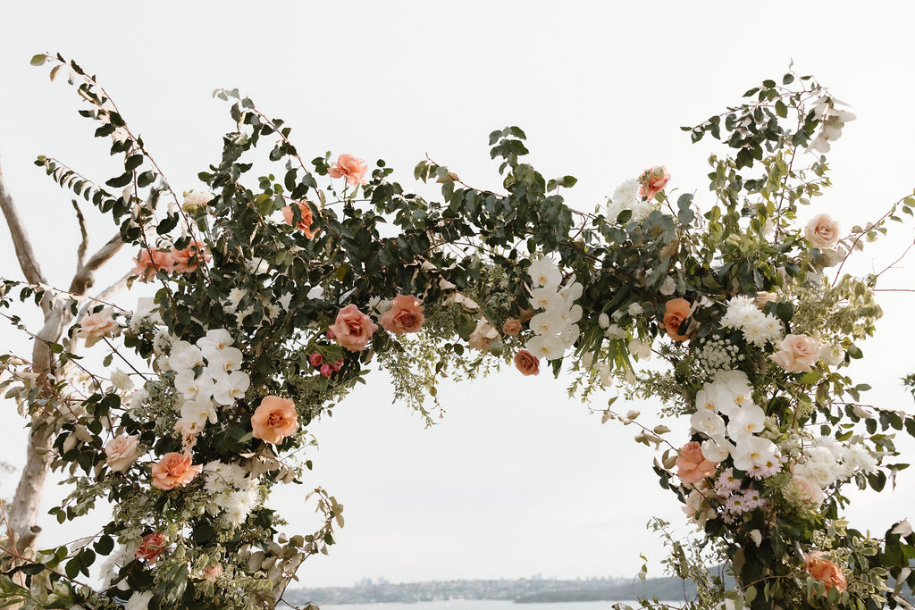 Floral wedding arch featuring cascading greenery, white orchids, blush and peach roses, and seasonal blooms, set outdoors with a soft coastal backdrop.