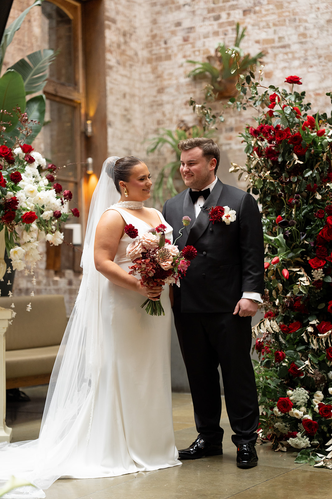 Modern romantic wedding at an industrial Sydney venue featuring a bride in a minimalist white gown and veil, groom in classic black tuxedo, surrounded by dramatic red and white floral arrangements with roses, dahlias, and gold foliage.