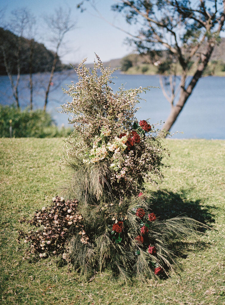 Foam-free floral ceremony tower with Australian native grasses, crimson roses and white flannel flowers on the Shoalhaven River lawn at Bundanon Trust, Southern Highlands wedding
